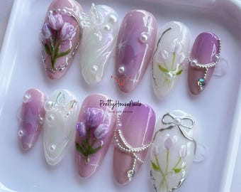 Könnte beinhalten: Set mit zehn falschen Nägeln in einem rosafarbenen und weißen Ombre-Design. Jeder Nagel zeigt ein anderes Blumenmuster mit Perlenakzenten und silbernen Verzierungen. Die Nägel sind in einer weißen Box angeordnet, auf deren Boden der Text "PrettyHouseNails LOVE LIFE DESIGN" gedruckt ist.