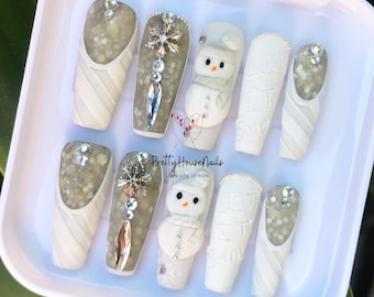 Pode incluir: Conjunto de dez pontas de unhas de inverno brancas e prateadas com desenhos de boneco de neve e flocos de neve. As pontas estão dispostas em uma caixa branca com o texto "PrettyHouseNails Love Life Design".