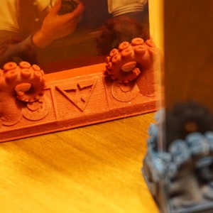 Pode incluir: Um porta-retratos vermelho impresso em 3D com um design de polvo na parte inferior. O quadro tem um inserto de pl&aacute;stico transparente e um s&iacute;mbolo de tri&acirc;ngulo na parte frontal.
