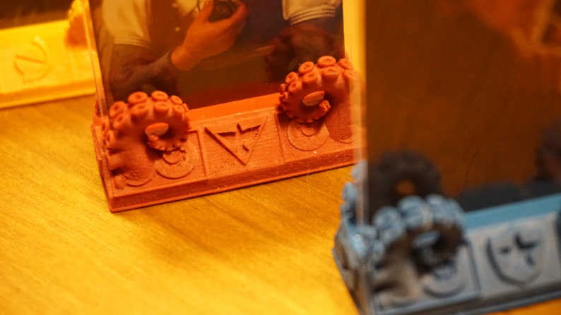 Pode incluir: Um porta-retratos vermelho impresso em 3D com um design de polvo na parte inferior. O quadro tem um inserto de pl&aacute;stico transparente e um s&iacute;mbolo de tri&acirc;ngulo na parte frontal.