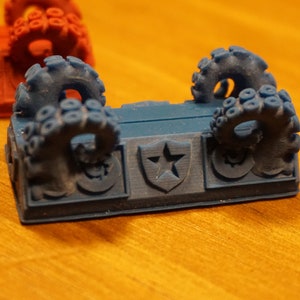 Pode incluir: Uma pe&ccedil;a de jogo impressa em 3D azul com dois tent&aacute;culos de polvo de cada lado. A pe&ccedil;a tem uma estrela em um design de escudo no centro.