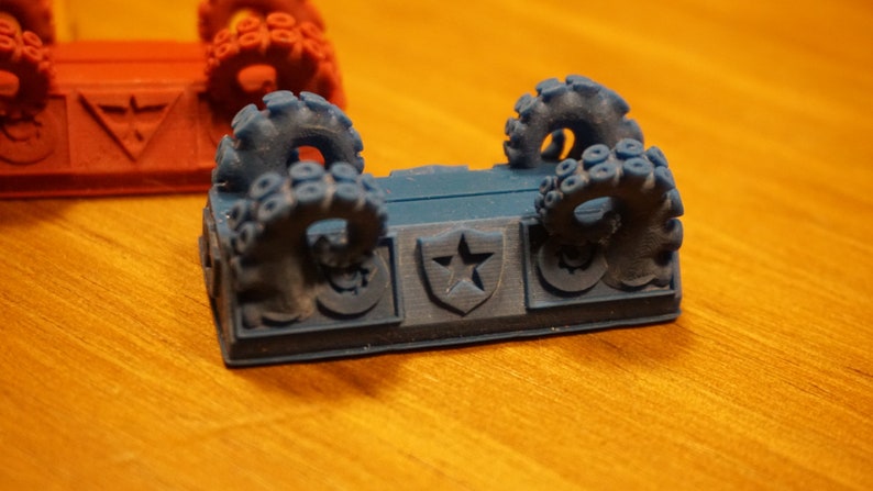 Pode incluir: Uma pe&ccedil;a de jogo impressa em 3D azul com dois tent&aacute;culos de polvo de cada lado. A pe&ccedil;a tem uma estrela em um design de escudo no centro.