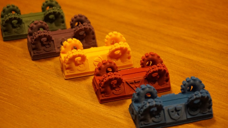 Pode incluir: Seis ba&uacute;s em miniatura coloridos com al&ccedil;as em forma de polvo. Os ba&uacute;s est&atilde;o dispostos em fila da esquerda para a direita, com as cores que v&atilde;o do verde escuro ao azul. Cada ba&uacute; tem um design diferente na parte frontal.