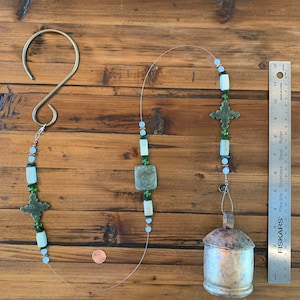 May include: A green and white beaded wind chime with a silver bell hanging from a hook. The chime features green and white beads, green cross-shaped charms, and square green stones.