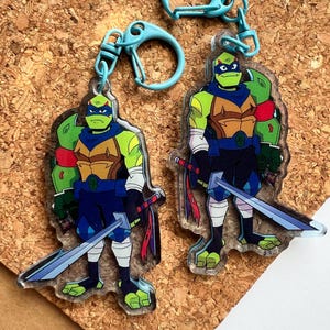 Puede incluir: Dos llaveros de acrílico con un personaje de tortuga de dibujos animados con piel verde, máscara azul y peto marrón. El personaje viste pantalones cortos azul oscuro, vendas blancas en las piernas y sostiene una espada larga plateada.