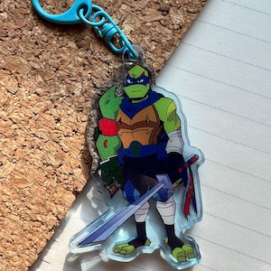 May include: A clear acrylic keyring featuring a cartoon character in green, blue, and brown. The character is holding a sword and has a blue mask. The keyring has a blue clasp.