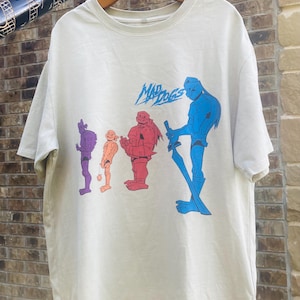 Könnte beinhalten: Ein beiges Kurzarm-T-Shirt mit einem Grafikdruck von vier stilisierten Figuren in Lila, Orange, Rot und Blau. Über den Figuren ist der Text "MAD DOGS" in einer blauen, gezackten Schriftart aufgedruckt. Das Shirt hat einen Rundhalsausschnitt.