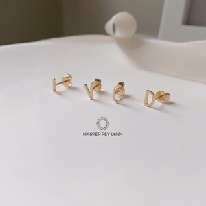 May include: Four gold-colored initial earrings, spelling out the word "LOVE". The earrings are on a white surface with the text "HARPER REY LYNN" printed below.
