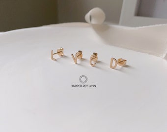 May include: Four gold-colored initial earrings, spelling out the word "LOVE". The earrings are on a white surface with the text "HARPER REY LYNN" printed below.