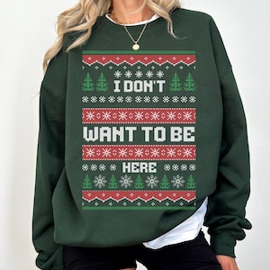 K&ouml;nnte beinhalten: Dunkelgr&uuml;nes Sweatshirt mit festlichem Design. Das Design enth&auml;lt den Text "I DON'T WANT TO BE HERE" in Wei&szlig;, umgeben von roten, gr&uuml;nen und wei&szlig;en Schneeflocken- und Baummustern. Das Sweatshirt hat einen Rundhalsausschnitt und lange &Auml;rmel.