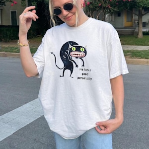 May include: White t-shirt featuring a black cartoon monster with green eyes and the text "He has a good personality." The graphic tee is made of a soft, comfortable material.