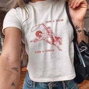 May include: A white t-shirt featuring a red illustration of a cowboy on a horse, with the words "save a horse" and "ride a cowboy". The shirt is worn with blue denim jeans.
