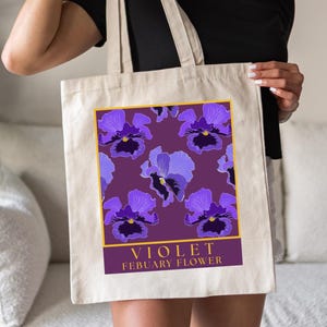 Może przedstawiać: Biała torba typu tote z płótna z fioletowym i złotym wzorem przedstawiającym kwiaty fiołków. Na torbie nadrukowany jest tekst "VIOLET FEBRUARY FLOWER".