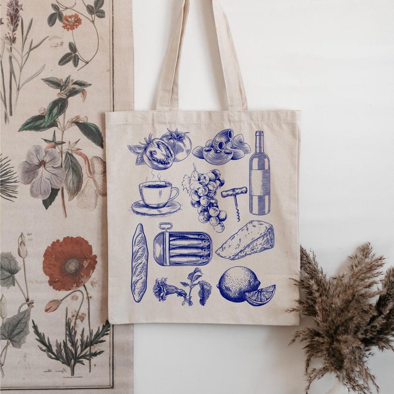 K&ouml;nnte beinhalten: Eine wei&szlig;e Einkaufstasche mit einer blauen Illustration verschiedener Lebensmittel, darunter ein Baguette, Trauben, K&auml;se, eine Flasche Wein, Tomaten und eine Dose Sardinen.