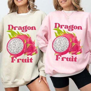 May include: Two women wearing sweatshirts with a graphic of a dragon fruit and the text "Dragon Fruit". The sweatshirt on the left is sand colored and the sweatshirt on the right is light pink.
