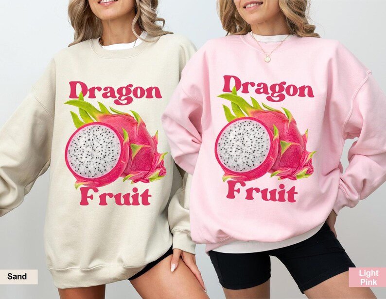 May include: Two women wearing sweatshirts with a graphic of a dragon fruit and the text "Dragon Fruit". The sweatshirt on the left is sand colored and the sweatshirt on the right is light pink.