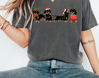 May include: A gray t-shirt with a graphic of four black dachshund dogs wearing Christmas hats and accessories. The dogs are lined up in a row and are decorated with Christmas lights.