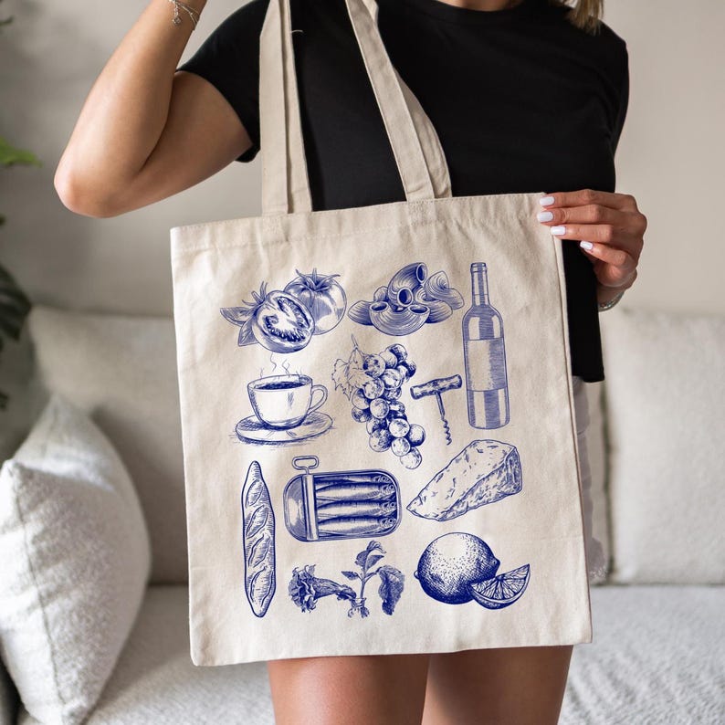 K&ouml;nnte beinhalten: Eine wei&szlig;e Canvas-Tragetasche mit blauen Illustrationen von verschiedenen Lebensmitteln, darunter eine Flasche Wein, ein Laib Brot, eine Dose Sardinen, eine Traube, eine Zitrone, eine Tasse Kaffee, Tomaten und Pasta.