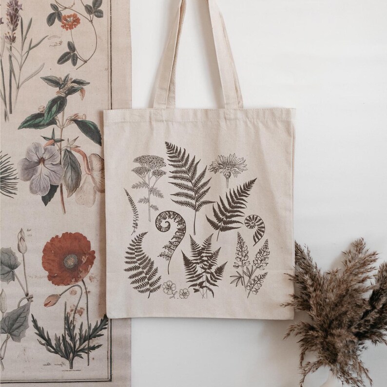 May include: A beige canvas tote bag with a black and white botanical print featuring ferns, flowers, and leaves. The print is reminiscent of vintage illustrations.