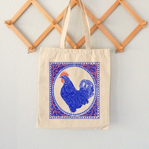 May include: A beige tote bag with a blue rooster illustration inside a red and blue patterned frame. The rooster is facing right and has a red comb.
