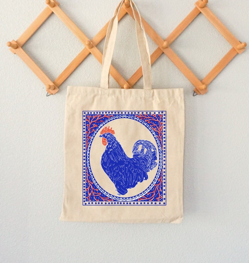 May include: A beige tote bag with a blue rooster illustration inside a red and blue patterned frame. The rooster is facing right and has a red comb.