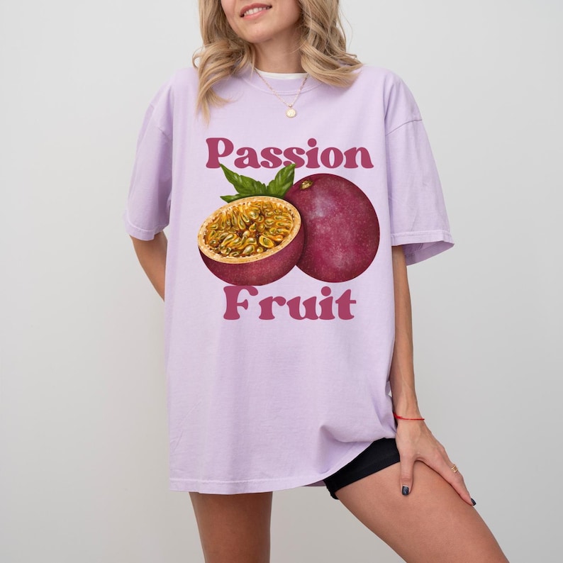 May include: A light purple t-shirt featuring a graphic of a passion fruit. The text "Passion Fruit" is printed in pink letters on the shirt.