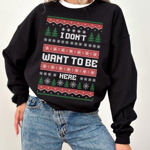K&ouml;nnte beinhalten: Schwarzes Sweatshirt mit dem Text "I DON'T WANT TO BE HERE" in Wei&szlig;, Rot und Gr&uuml;n, mit Schneeflocken- und Weihnachtsbaum-Motiven. Das Sweatshirt wird mit blauen Jeans kombiniert. Ein wei&szlig;es T-Shirt ist darunter sichtbar.