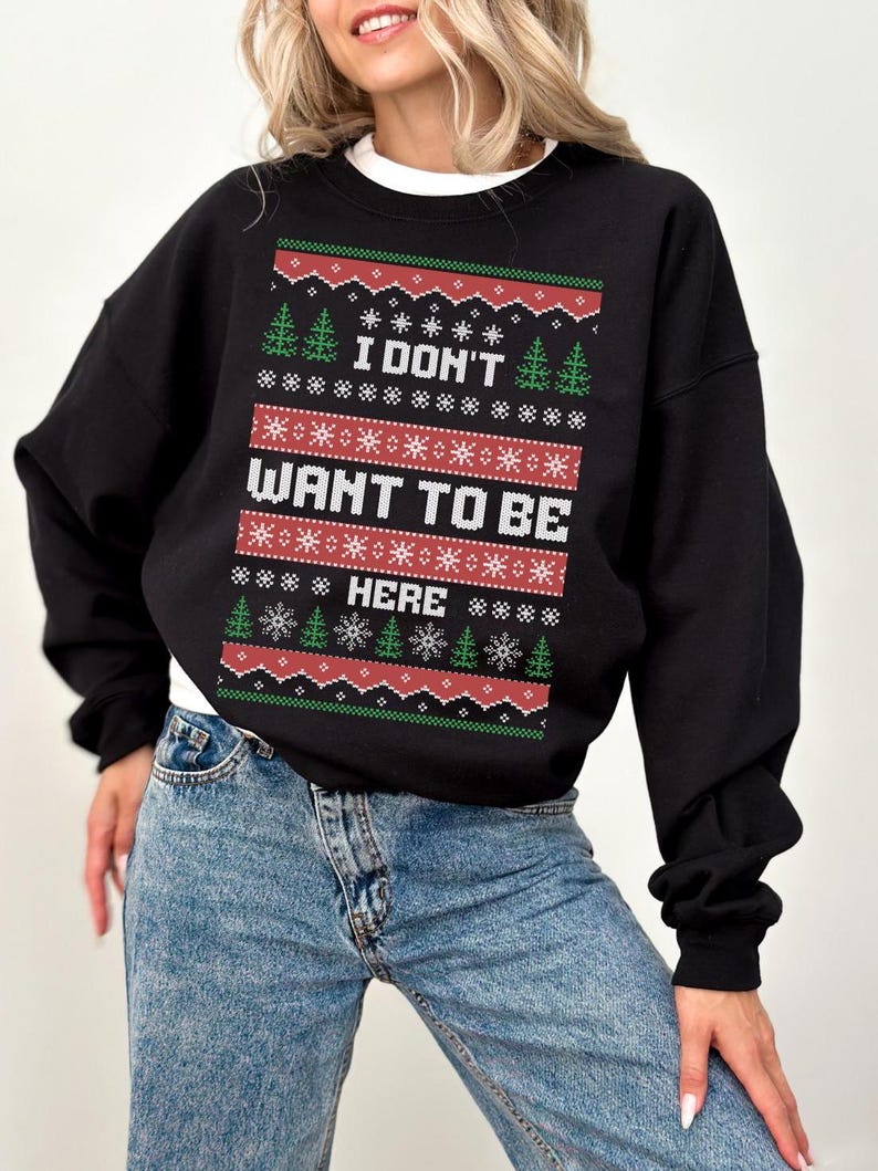 K&ouml;nnte beinhalten: Schwarzes Sweatshirt mit dem Text "I DON'T WANT TO BE HERE" in Wei&szlig;, Rot und Gr&uuml;n, mit Schneeflocken- und Weihnachtsbaum-Motiven. Das Sweatshirt wird mit blauen Jeans kombiniert. Ein wei&szlig;es T-Shirt ist darunter sichtbar.