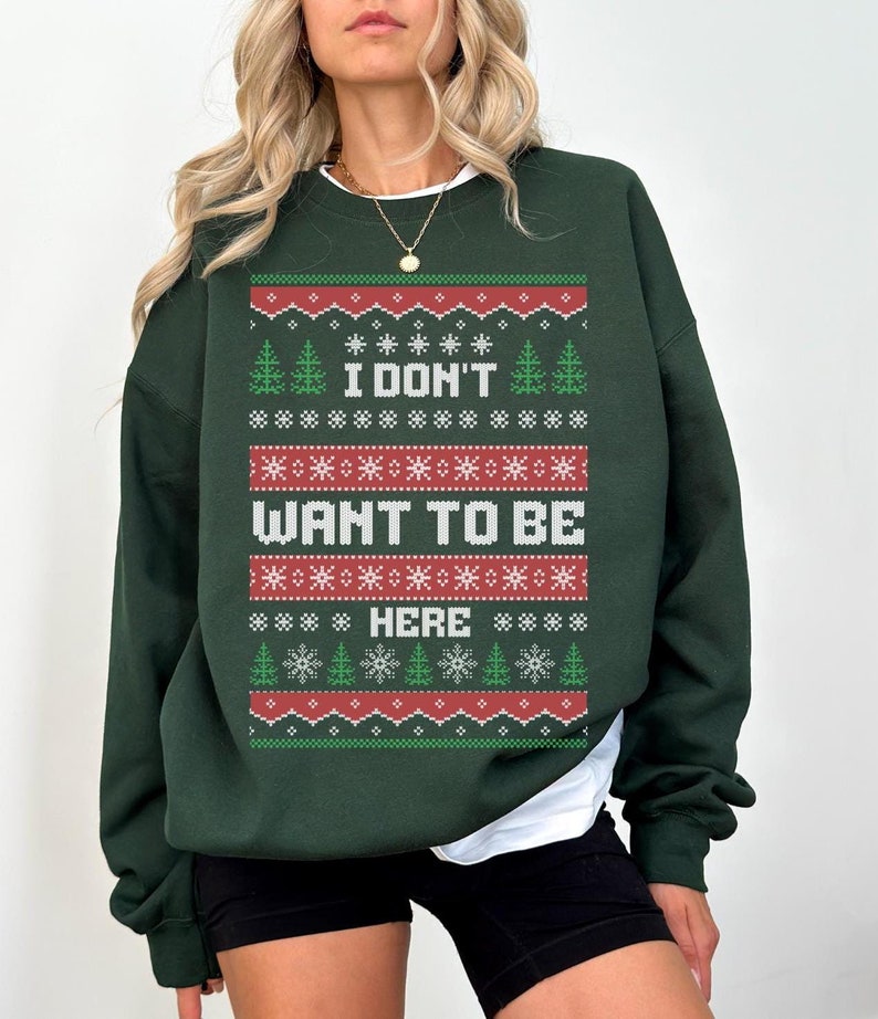 K&ouml;nnte beinhalten: Dunkelgr&uuml;nes Sweatshirt mit festlichem Design. Das Design enth&auml;lt den Text "I DON'T WANT TO BE HERE" in Wei&szlig;, umgeben von roten, gr&uuml;nen und wei&szlig;en Schneeflocken- und Baummustern. Das Sweatshirt hat einen Rundhalsausschnitt und lange &Auml;rmel.