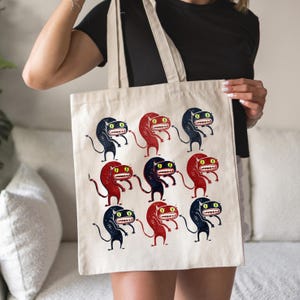May include: Beige canvas tote bag featuring a repeating pattern of cartoonish creatures in red and navy blue. Each creature has large eyes and a toothy grin. The bag has a simple, rectangular shape with long handles.