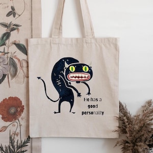 May include: Beige canvas tote bag featuring a cartoon illustration of a black creature with green eyes and a wide grin. The text "He has a good personality" is printed below the illustration.