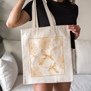 May include: A white canvas tote bag with a design of orange slices and leaves in a gold color.