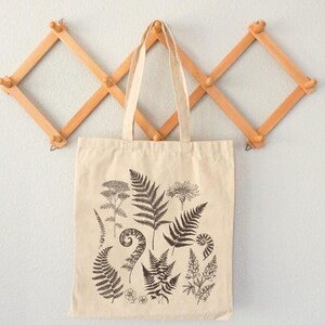 May include: A beige tote bag with a black and white print of various ferns and flowers. The bag is hanging on a wooden wall rack.