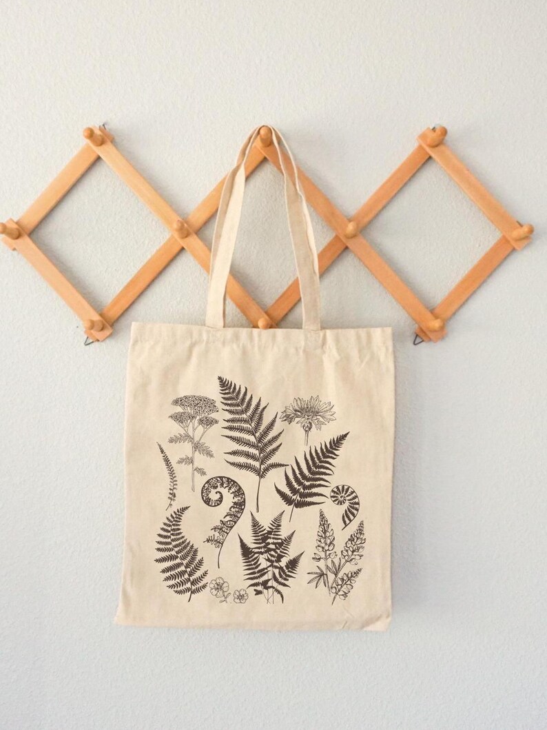 May include: A beige tote bag with a black and white print of various ferns and flowers. The bag is hanging on a wooden wall rack.