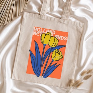 Może przedstawiać: Biała torba typu tote z pł&oacute;tna z graficznym wzorem ż&oacute;łtych tulipan&oacute;w na jasno pomarańczowym tle. Tekst "HOLLAND NETHERLANDS" jest nadrukowany na torbie czarnym kolorem.