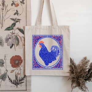 May include: A white tote bag with a blue rooster illustration inside a red and blue patterned frame. The rooster is blue with a red comb and wattle.