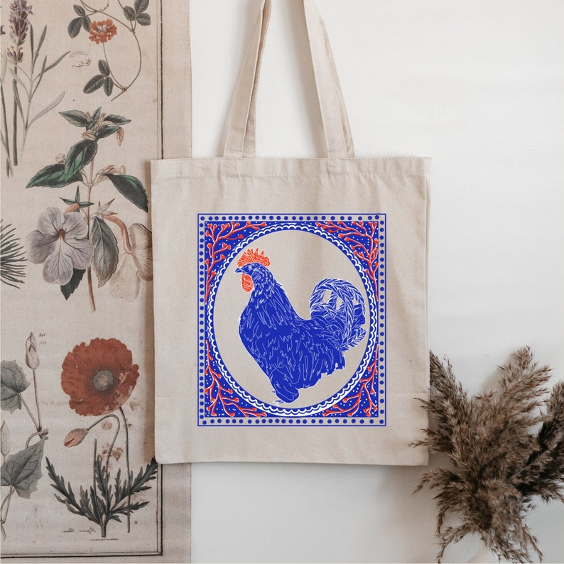 May include: A white tote bag with a blue rooster illustration inside a red and blue patterned frame. The rooster is blue with a red comb and wattle.