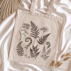 May include: A beige canvas tote bag with a black and white botanical print featuring ferns, flowers, and leaves. The print is centered on the bag and covers most of the surface.