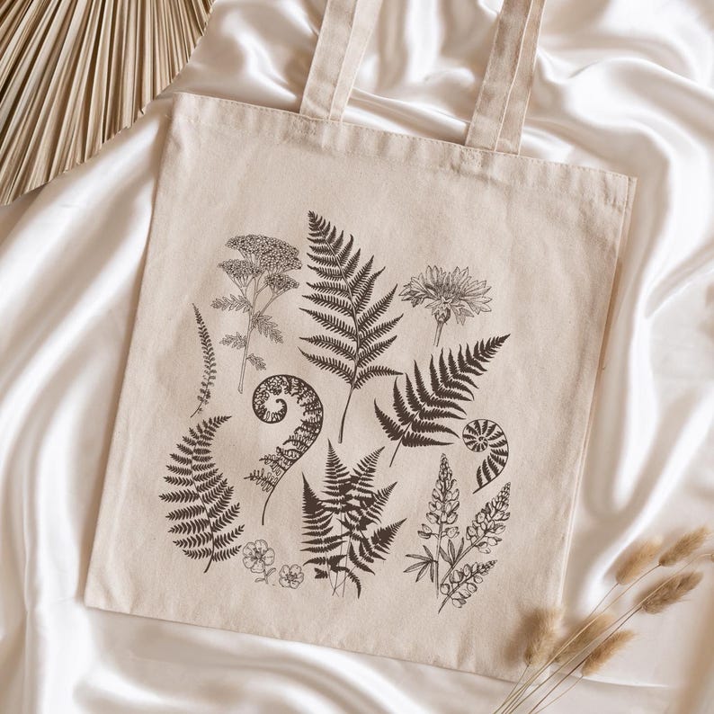 May include: A beige canvas tote bag with a black and white botanical print featuring ferns, flowers, and leaves. The print is centered on the bag and covers most of the surface.