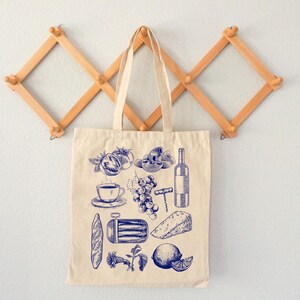 K&ouml;nnte beinhalten: Eine beige Canvas-Tragetasche mit blauen Illustrationen verschiedener Lebensmittel, darunter ein Baguette, Trauben, eine Flasche Wein, eine Zitrone, eine Tasse Kaffee, ein St&uuml;ck K&auml;se und eine Dose Sardinen.