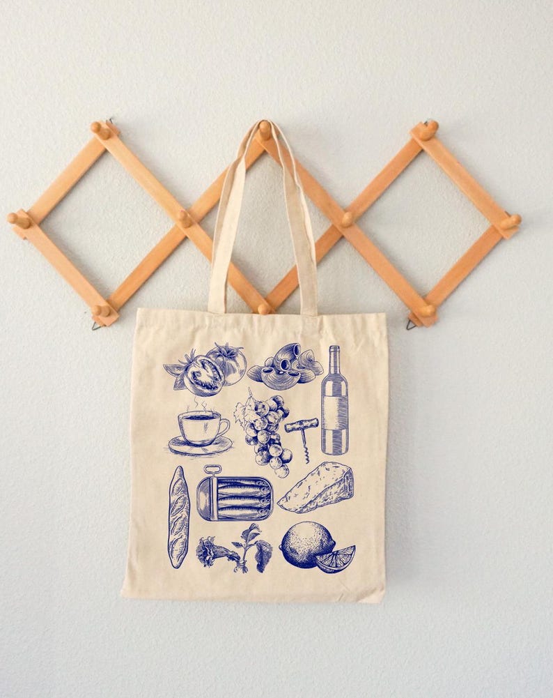 K&ouml;nnte beinhalten: Eine beige Canvas-Tragetasche mit blauen Illustrationen verschiedener Lebensmittel, darunter ein Baguette, Trauben, eine Flasche Wein, eine Zitrone, eine Tasse Kaffee, ein St&uuml;ck K&auml;se und eine Dose Sardinen.