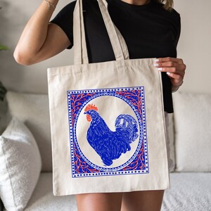 May include: A white canvas tote bag with a blue rooster illustration inside a red and blue patterned frame. The rooster is in a circle with a red and blue floral design around it.
