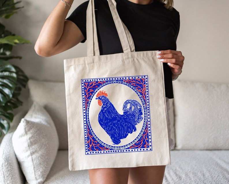 May include: A white canvas tote bag with a blue rooster illustration inside a red and blue patterned frame. The rooster is in a circle with a red and blue floral design around it.