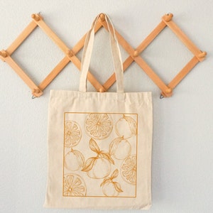May include: A beige canvas tote bag with a design of orange slices and whole oranges in a golden yellow color. The bag is hanging on a wooden rack.