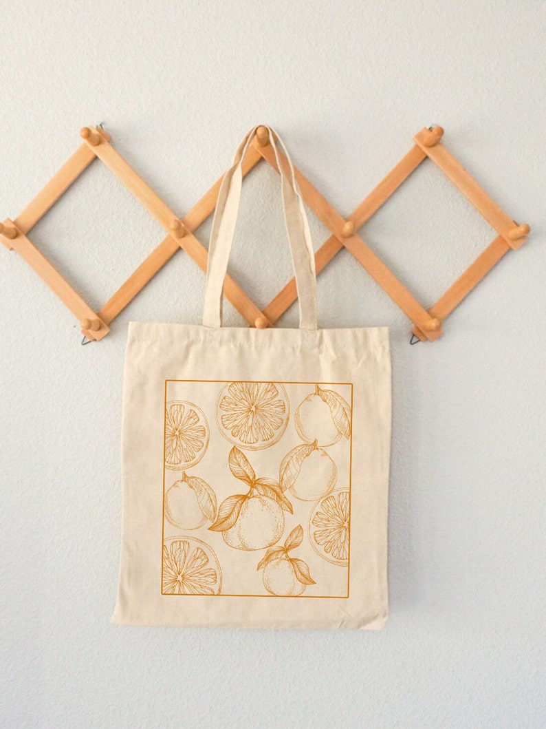 May include: A beige canvas tote bag with a design of orange slices and whole oranges in a golden yellow color. The bag is hanging on a wooden rack.