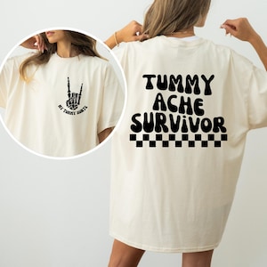 May include: A white t-shirt with a black graphic on the front that says "My Twenty Hurts" and a black graphic on the back that says "Tummy Ache Survivor" with a black and white checkered pattern below the text.