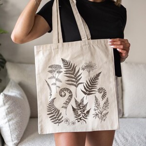 May include: A beige canvas tote bag with a black and white botanical print featuring ferns, flowers, and leaves. The print is centered on the bag and covers most of the front.