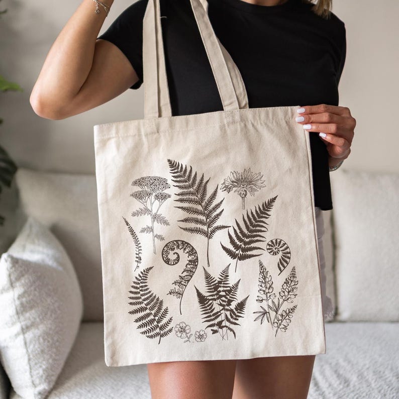 May include: A beige canvas tote bag with a black and white botanical print featuring ferns, flowers, and leaves. The print is centered on the bag and covers most of the front.
