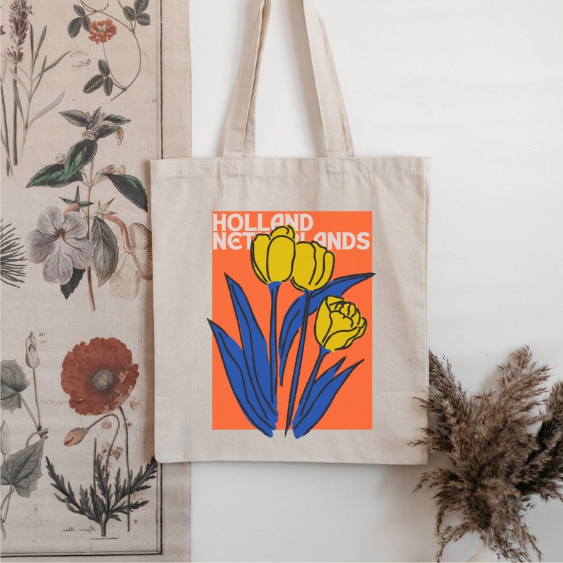 Może przedstawiać: Biała torba typu tote z kolorową ilustracją ż&oacute;łtych tulipan&oacute;w i niebieskich liści na pomarańczowym tle. Tekst "HOLLAND NETHERLANDS" jest nadrukowany czarnym kolorem u g&oacute;ry ilustracji.