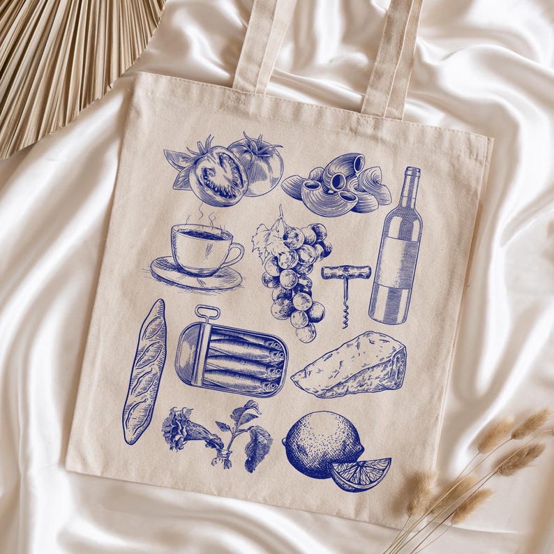 K&ouml;nnte beinhalten: Eine beige Stofftasche mit einer blauen Illustration verschiedener Lebensmittel, darunter Tomaten, Trauben, Pasta, ein Baguette, eine Dose Sardinen, K&auml;se, eine Zitrone, eine Tasse Kaffee, eine Weinflasche und ein Korkenzieher.